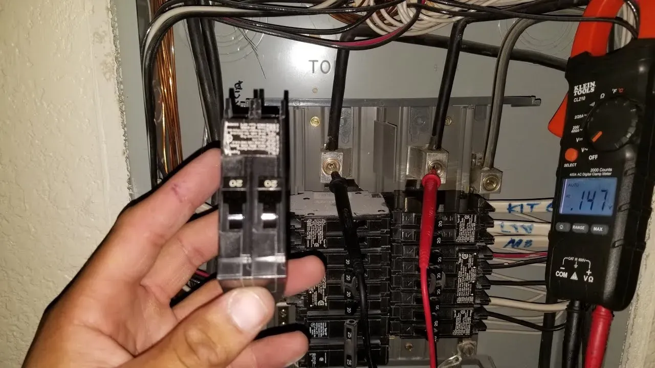 Electrician holding a 20 amp double-pole breaker in front of an open panel while a clamp meter tests the main lugs