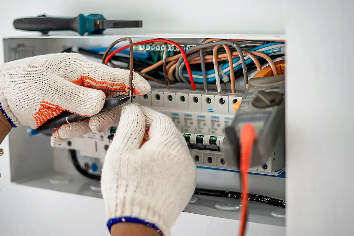 Gloved hands working on wires inside a home breaker panel with a multimeter beside it