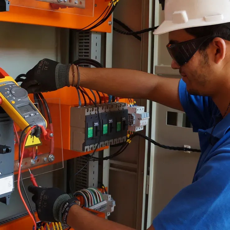 Electrician in a hard hat and safety glasses wiring breakers in an electrical panel with a clamp meter