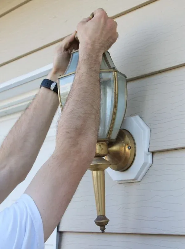 Hands removing an old brass porch light from the siding of a house