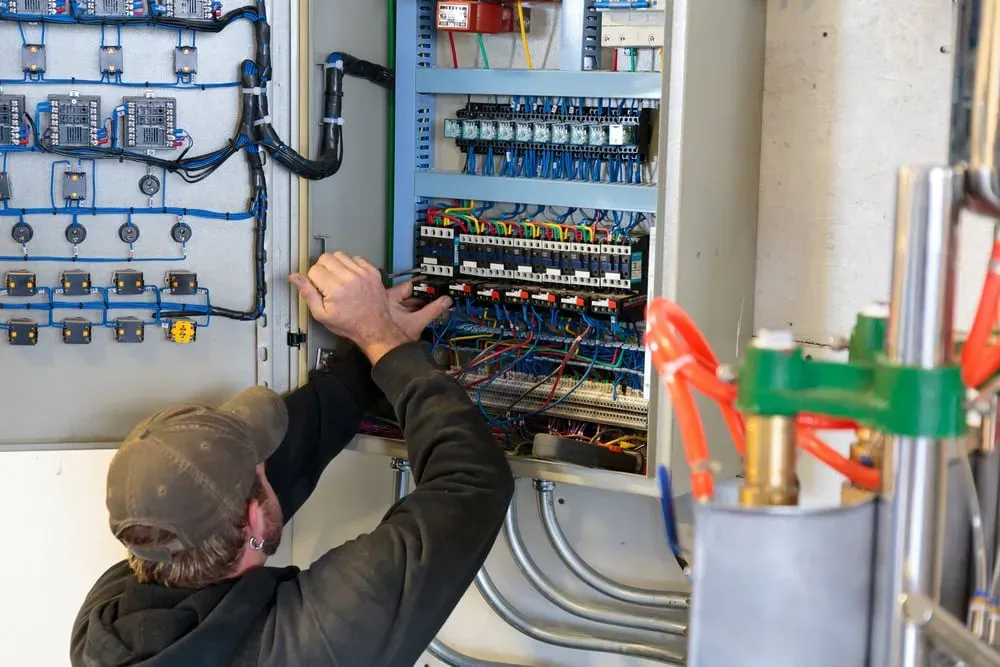 Electrician in a cap working on wiring inside a large open control panel