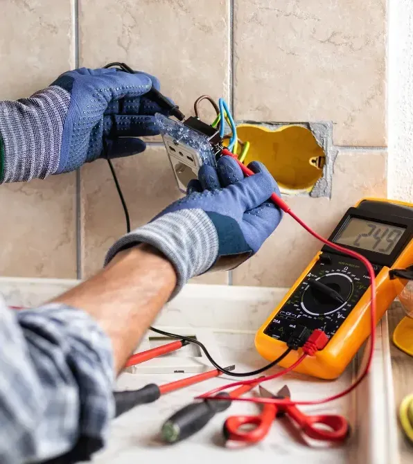 Electrician in gloves testing an outlet with a multimeter before wiring it into a wall box